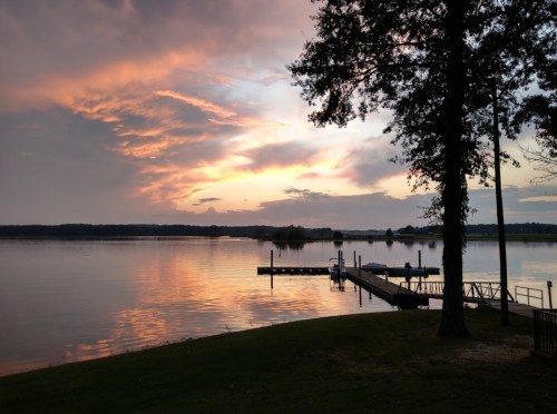 A serene lake at sunset, with colorful clouds reflecting on the water and a dock extending into the scene.