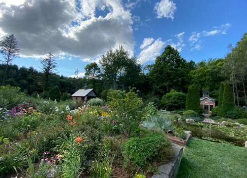A vibrant garden with colorful flowers, a pond, and a quaint house surrounded by lush trees under a blue sky.