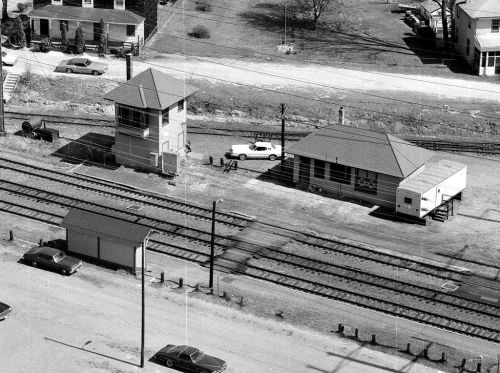 Aerial view of train tracks with a control tower, a building, and parked cars in a rural setting.