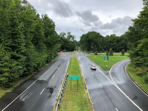 A view of a wet road intersection surrounded by greenery under a cloudy sky. Road signs indicate directions.