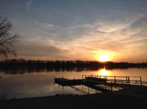 A serene sunset over a calm lake, with a dock extending into the water and trees silhouetted in the background.