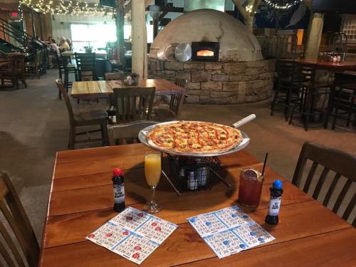 A wooden table with a large pizza, drinks, and game cards, set in a cozy restaurant with a stone pizza oven in the background.