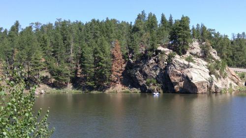 A serene lake surrounded by lush green trees and a rocky shoreline under a clear blue sky.