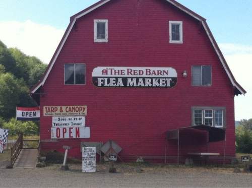 A red barn with signs for "The Red Barn Flea Market" and "OPEN," surrounded by a gravel area and greenery.