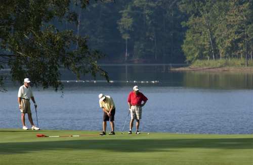 Three golfers on a green by a lake, with trees in the background and one player preparing to putt.