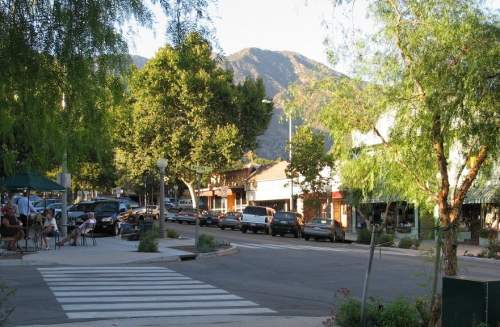 A quiet street scene with trees, parked cars, and shops, set against a backdrop of mountains in the distance.