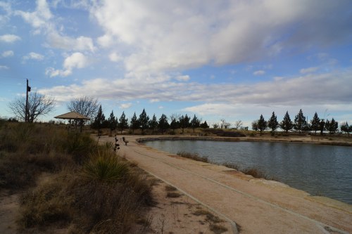A serene landscape featuring a pond, grassy shore, and trees under a cloudy sky.