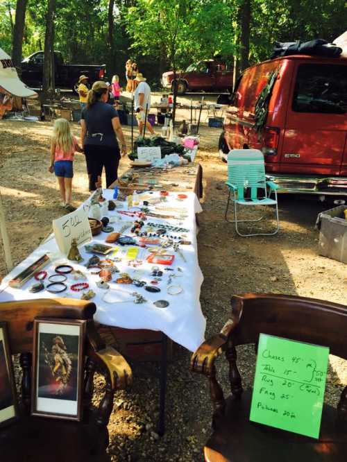 A bustling outdoor market with tables displaying jewelry and crafts, surrounded by trees and people browsing.