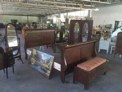 A spacious warehouse filled with various wooden furniture pieces, including beds, mirrors, and a painting.