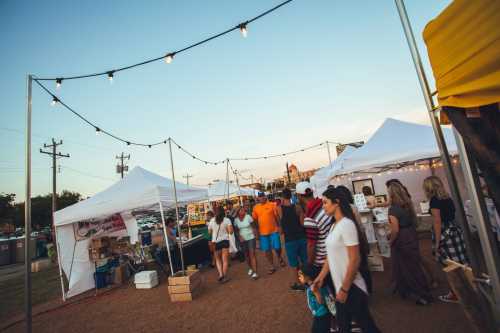 A lively outdoor market scene with people walking among white tents and string lights at sunset.