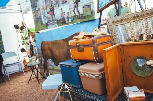 A collection of vintage suitcases and antiques displayed at an outdoor market, with a mural of a cow in the background.