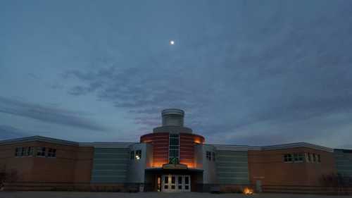 A modern building at dusk, illuminated with soft lights, under a cloudy sky with a visible moon.