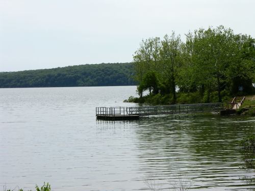 A calm lake scene with a wooden dock and lush green trees along the shoreline under a cloudy sky.