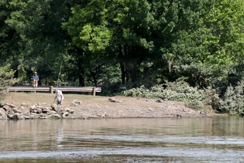 A person fishing by a riverbank while another child stands on a nearby dock surrounded by trees.
