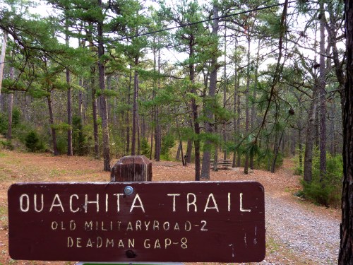 Sign for Ouachita Trail with a wooded path in the background, surrounded by tall pine trees and autumn foliage.