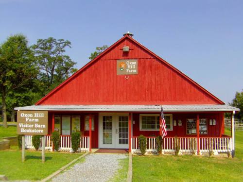 Red barn-style building with a sign for Oxon Hill Farm, featuring a porch and American flag in front. Clear blue sky.