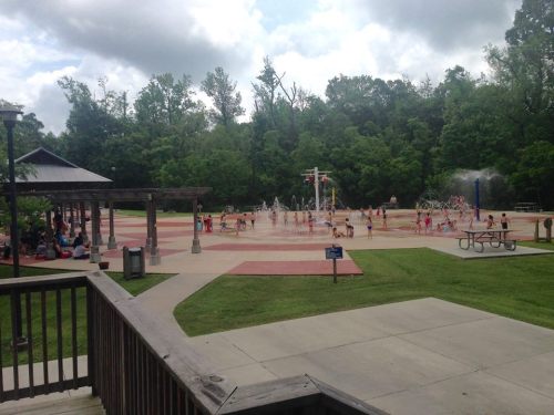 A splash pad with children playing in water, surrounded by trees and picnic areas on a cloudy day.