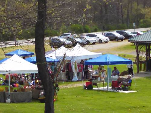 A vibrant outdoor market with colorful tents, vendors, and visitors enjoying the sunny day in a grassy area.