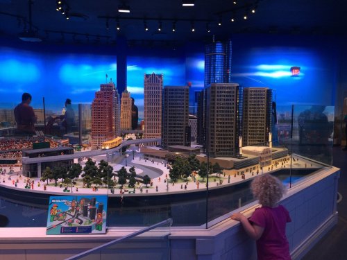 A child observes a detailed miniature cityscape with skyscrapers and trees in a brightly lit exhibit.