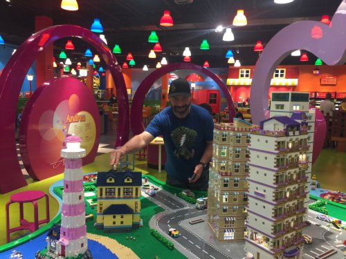 A man interacts with colorful LEGO buildings in a vibrant play area filled with bright lights and playful decor.