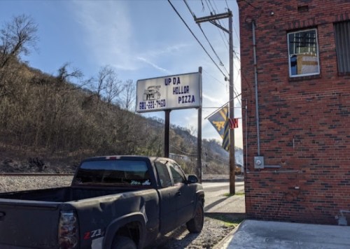 A black pickup truck parked near a sign for "Up Da Hollor Pizza" beside a brick building and a hillside.