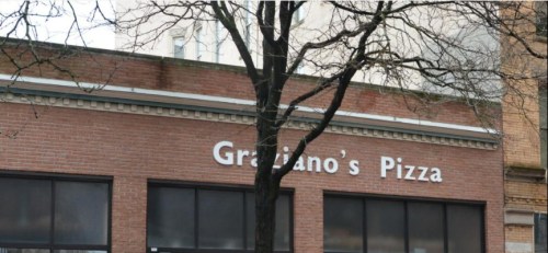 Sign for "Graziano's Pizza" on a brick building, partially obscured by a tree.