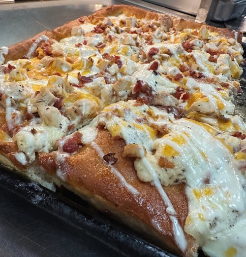A close-up of a freshly baked pizza topped with chicken, bacon, cheese, and drizzled with sauce on a dark baking tray.