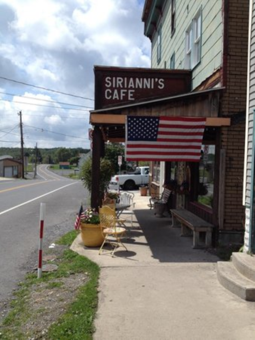 A quaint café with a sign reading "Sirianni's Café," featuring an American flag and outdoor seating along a road.