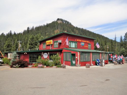 A red wooden store surrounded by trees, with flags and flowers, set against a mountain backdrop.
