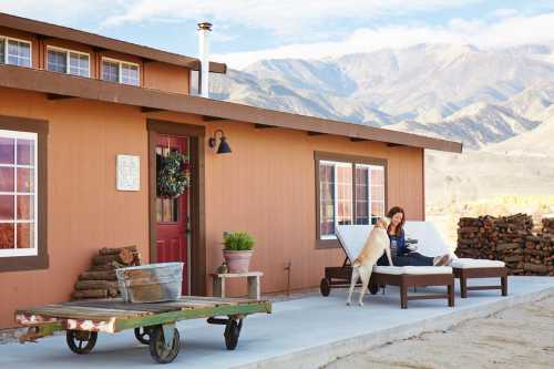 A woman sits on a lounge chair outside a rustic house, with a dog nearby and mountains in the background.