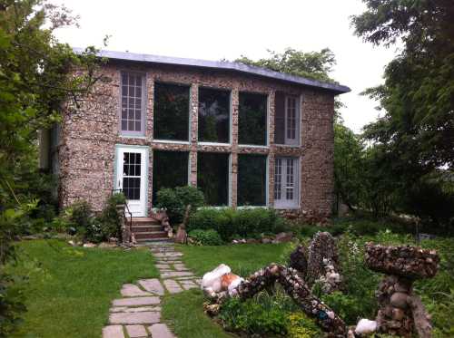 A unique stone house with large windows, surrounded by greenery and a stone pathway in the front yard.