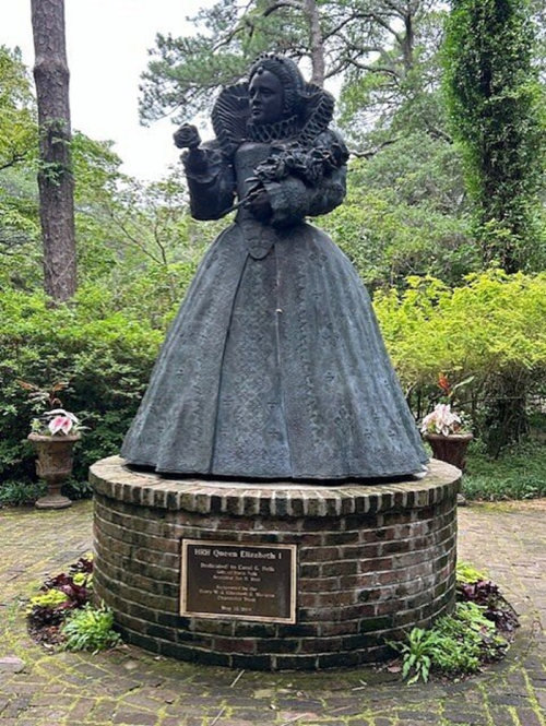 Statue of Queen Elizabeth I in a garden, wearing a detailed gown, standing on a brick pedestal with a plaque.