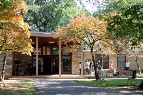 A modern building surrounded by trees, with people setting up equipment outside on a sunny day.