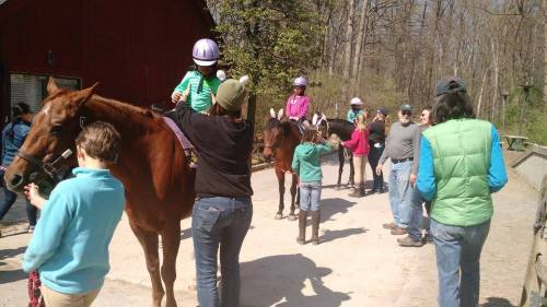 A group of people, including children on horseback, gather near a stable in a wooded area on a sunny day.