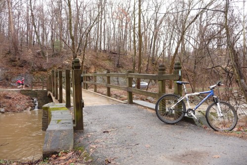 A mountain bike rests on a gravel path beside a wooden bridge over a small stream in a wooded area.