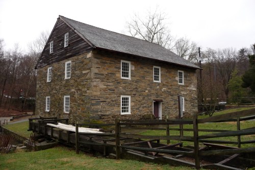 A rustic stone building with a wooden roof, surrounded by greenery and a wooden fence, on a cloudy day.