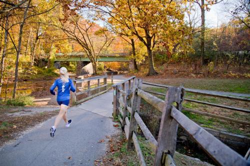 A person runs along a path surrounded by autumn trees and a wooden fence, with a bridge in the background.