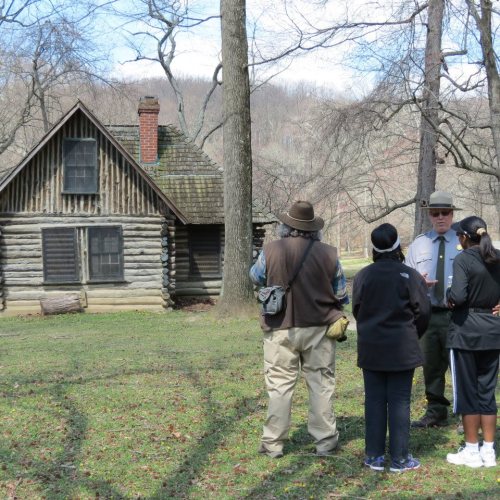 A park ranger talks to two visitors near a log cabin in a wooded area, with trees and hills in the background.