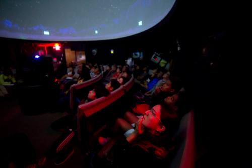 A group of children sitting in a dark theater, gazing up at a starry projection on the dome ceiling.