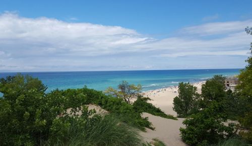 A scenic view of a beach with turquoise water, sandy shore, and lush greenery under a partly cloudy sky.