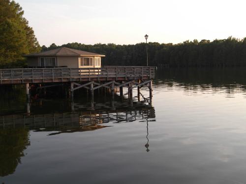 A wooden pier extends over calm water, leading to a small, round building surrounded by trees at sunset.