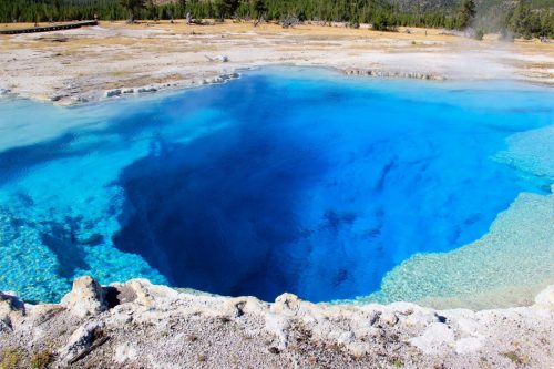 Vibrant blue hot spring surrounded by rocky edges and a natural landscape in Yellowstone National Park.