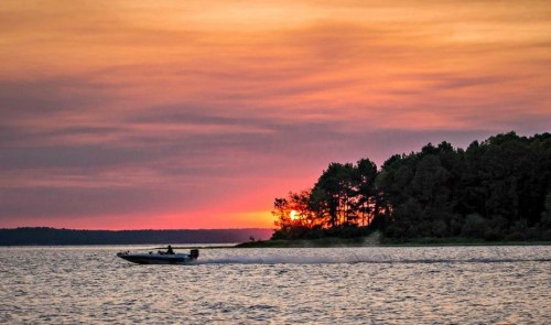 A boat glides across a lake at sunset, with trees silhouetted against a colorful sky.