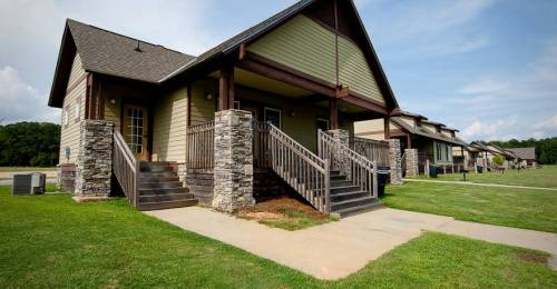 A modern house with stone accents and wooden stairs, set on a grassy area under a blue sky.