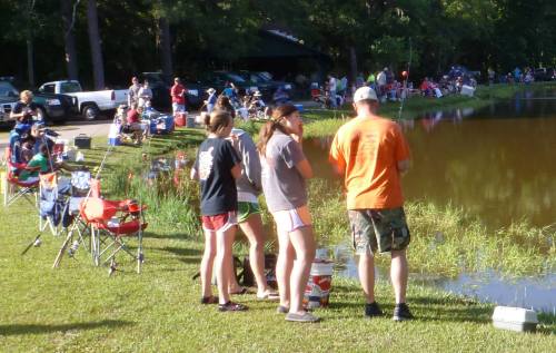 A group of people fishing by a pond, with chairs set up and a crowd in the background enjoying the outdoors.