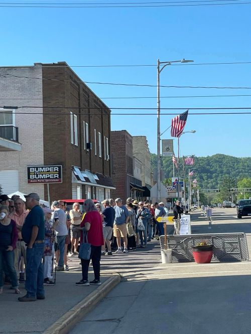 A long line of people waiting outside shops on a sunny day, with American flags lining the street.
