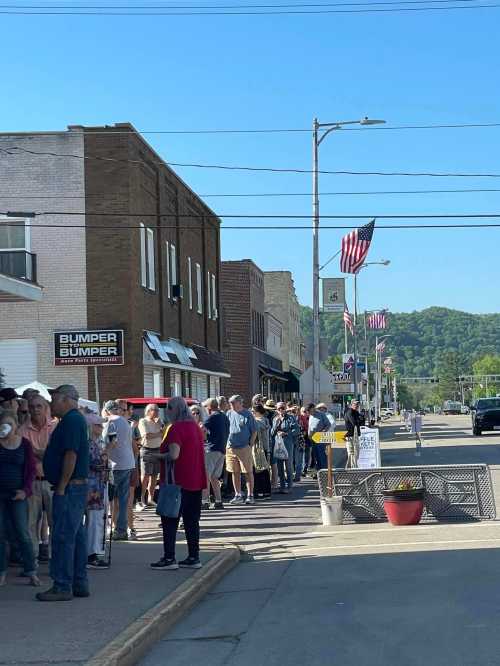 A long line of people waiting outside shops on a sunny day, with American flags lining the street.