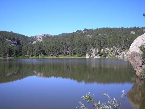A serene lake surrounded by lush green trees and rocky hills under a clear blue sky. Reflections shimmer on the water.
