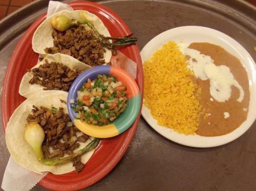 A plate of tacos with grilled meat and onions, accompanied by rice, refried beans, and a side of pico de gallo.