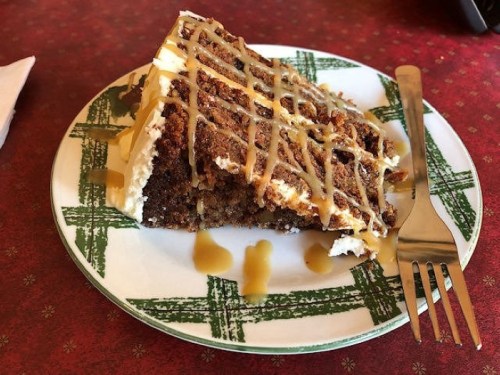 A slice of cake with cream frosting and caramel drizzle on a decorative plate, accompanied by a fork.